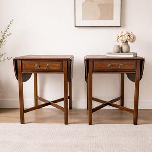 May include: Two dark brown wooden side tables with drop-leaf sides and a drawer. Each table has a brass handle on the drawer and a cross-legged base. The tables are placed on a light-colored rug.