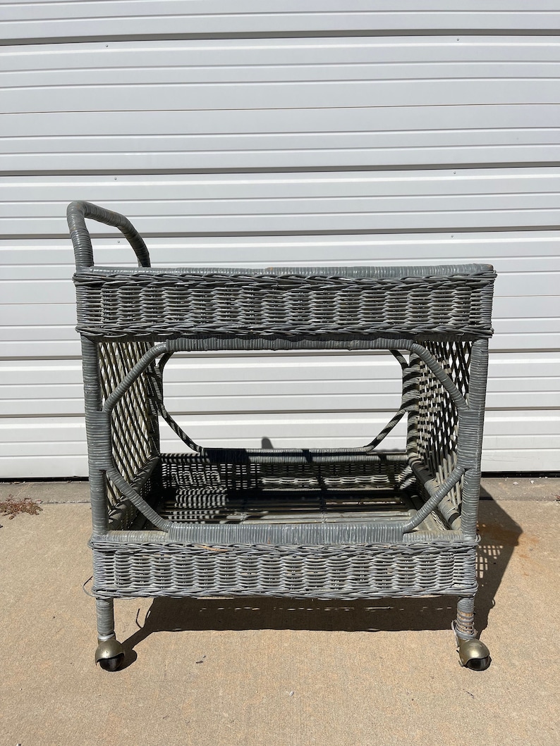 May include: A gray wicker serving cart with two shelves and wheels. The cart is made of woven wicker and has a rustic, vintage look.