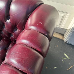 May include: Close-up of a burgundy leather sofa arm with a tufted design. The leather has a glossy finish and shows subtle variations in color. The armrest is well-padded and has a classic, elegant appearance.