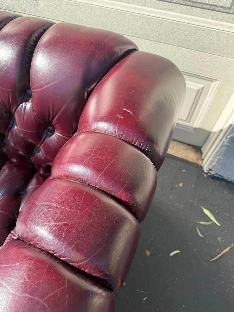 May include: Close-up of a burgundy leather sofa arm with a tufted design. The leather has a glossy finish and shows subtle variations in color. The armrest is well-padded and has a classic, elegant appearance.
