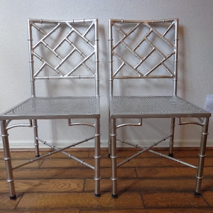 May include: Two silver chairs with bamboo-style frames. The chairs feature a lattice design on the backrests and woven seats. The legs are curved, and the chairs are on a wooden floor.