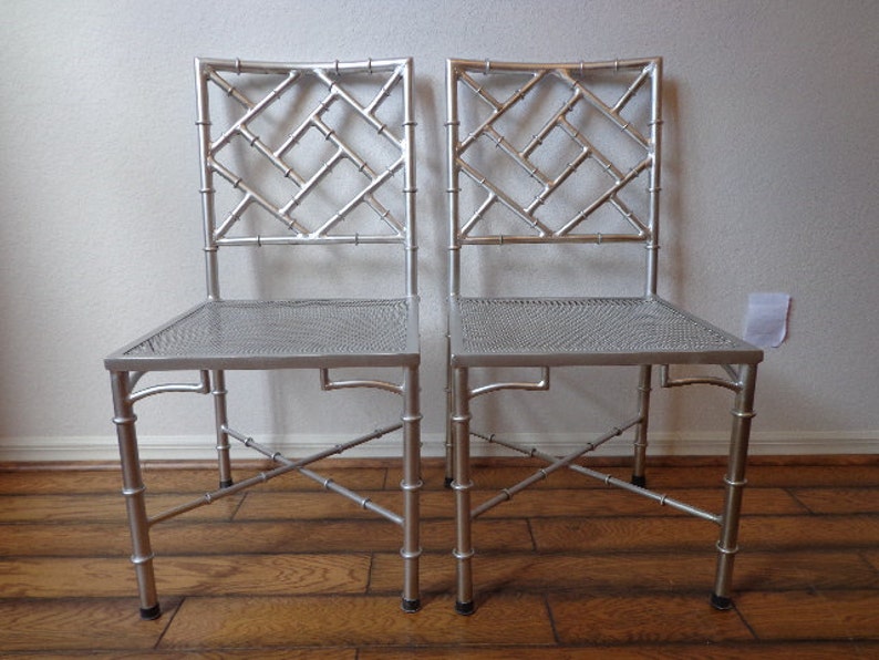 May include: Two silver chairs with bamboo-style frames. The chairs feature a lattice design on the backrests and woven seats. The legs are curved, and the chairs are on a wooden floor.