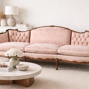 May include: A vintage-style, three-seat sofa with a light pink floral patterned fabric and dark wood trim. The sofa features tufted detailing on the armrests and back. A round marble coffee table and a vase of white flowers are in front of the sofa.