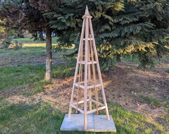 May include: A wooden pyramid-shaped trellis with a pointed top. The trellis is made of natural wood and has a light brown color. It is standing on a concrete patio.