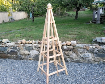 May include: A wooden garden trellis with a square base and a pointed top. The trellis is made of natural wood and has a light brown color. It is designed to support climbing plants.