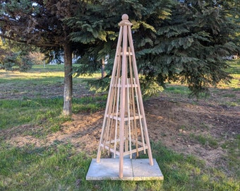 May include: A wooden garden trellis in the shape of a pyramid with a pointed top. The trellis is made of natural wood and has a light brown color. It is standing on a square concrete base in a grassy area.