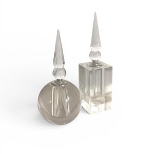 May include: Two clear glass perfume bottles. One is round, the other is square. Both have a clear, pointed stopper. The round bottle is a smoky gray color. The square bottle is clear. The bottles are on a white background.
