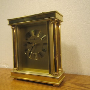 May include: A gold-toned mantel clock with Roman numeral markings on the face. The clock has a rectangular shape with decorative columns on the sides.