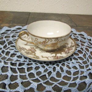 May include: A white teacup and saucer set with gold trim and floral and bird designs. The cup has a gold handle. The set is on a light blue crocheted doily. The teacup and saucer are likely porcelain.