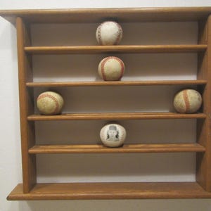 May include: A wooden baseball display shelf with five baseballs. The shelf is made of light brown wood and has four horizontal shelves. Four baseballs are standard, and one has a portrait. The baseballs are white with red stitching.