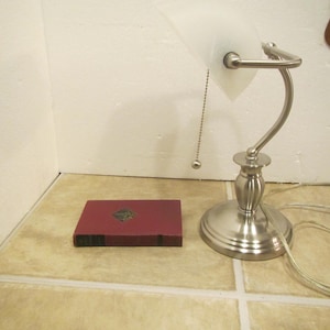 May include: A brushed nickel desk lamp with a frosted glass shade and a pull chain. A small, maroon book with gold accents is next to the lamp. The lamp has a curved arm and a round base.
