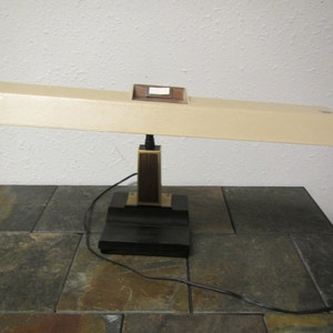 May include: A vintage beige desk lamp with a black base and a brown wooden accent. The lamp has a rectangular shade and a switch on the side.