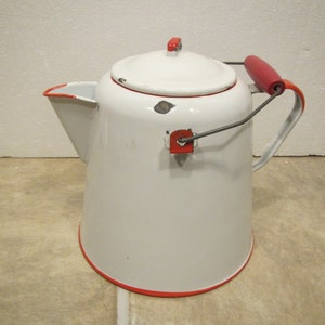 May include: A white enamelware coffee pot with a red trim and handle. The pot has a lid with a red handle and a spout for pouring. The handle is red and the pot has a metal handle for carrying. The pot is likely vintage.