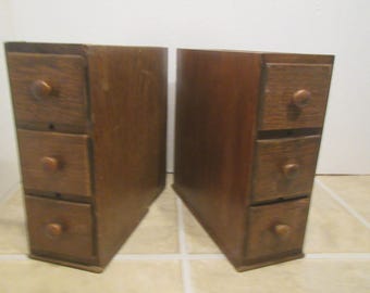 May include: Two matching wooden drawer units. Each unit has three drawers with round wooden knobs. The wood has a warm, medium brown tone. The units are side by side, with a white wall in the background.