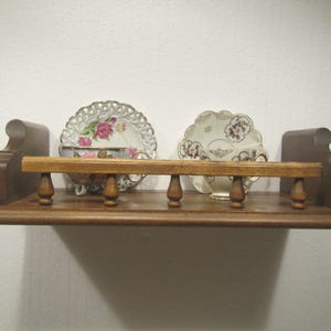 May include: A wooden shelf with a decorative design, holding two antique teacups and saucers. The shelf is brown with a decorative front rail and end supports. The teacups and saucers have floral patterns.