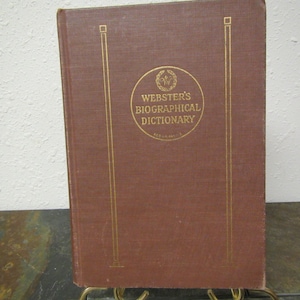 May include: A brown hardcover book with gold lettering on the spine and front cover. The title is "Webster's Biographical Dictionary".