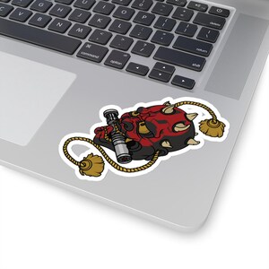 May include: A red and black cartoon character with horns, holding a lightsaber, is depicted on a white sticker with a rope and tassels. The sticker is placed on a silver laptop.