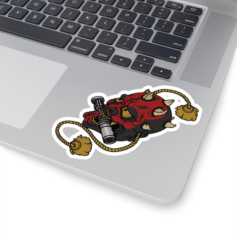 May include: A red and black cartoon character with horns, holding a lightsaber, is depicted on a white sticker with a rope and tassels. The sticker is placed on a silver laptop.