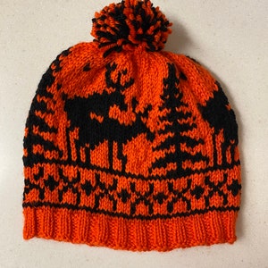 May include: An orange and black knit hat with a pom-pom. The hat features a pattern of trees and deer in black against an orange background. The brim is a solid orange color.