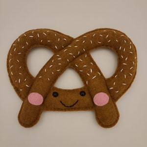 May include: A brown felt pretzel with white stitching and a smiling face. The pretzel has pink felt cheeks.