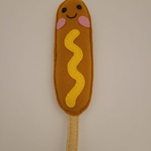 May include: A felt corndog toy with a smiling face, yellow mustard, and a brown stick.