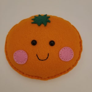Op de afbeelding: Een oranje van vilt met een groen blaadje erop, een glimlachend gezicht en roze wangen.