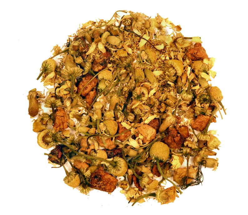 May include: A close-up shot of a pile of dried herbal tea blend. The blend features a mix of yellow chamomile flowers, small brown pieces, and green stems, all against a white background. The tea appears to be loose leaf.