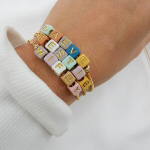 May include: A bracelet with colorful beads that spell out the names "Maeve", "West", "Wife", and "Lexy". The beads are made of a smooth, matte material and have gold lettering.
