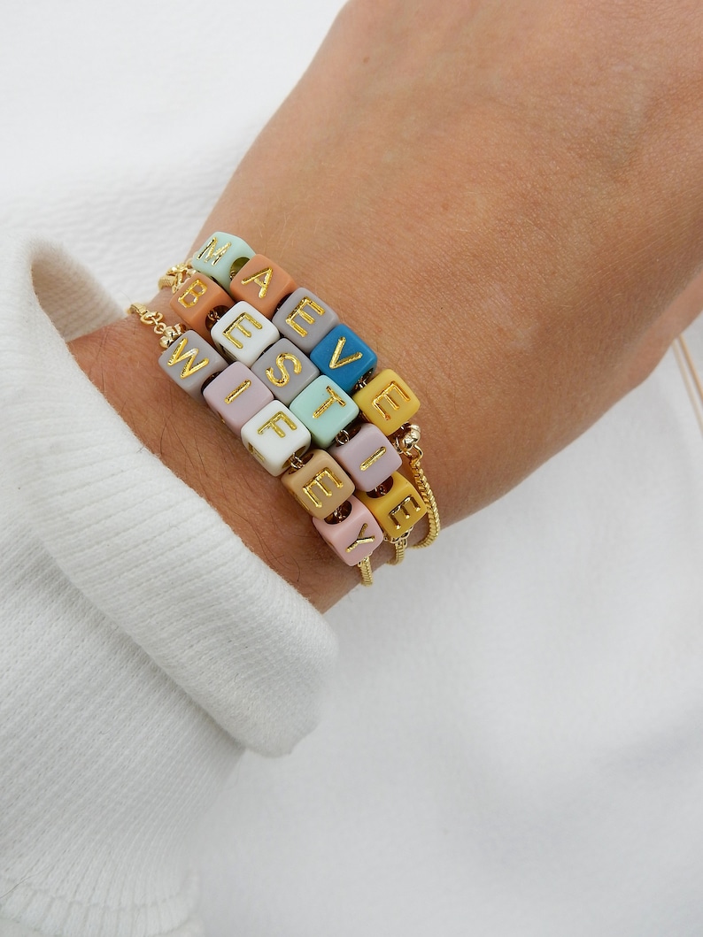 May include: A bracelet with colorful beads that spell out the names "Maeve", "West", "Wife", and "Lexy". The beads are made of a smooth, matte material and have gold lettering.
