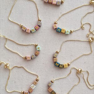 May include: A set of nine gold chain bracelets with colorful beads spelling out different names. The names include MOMMY, ZAMA, NANA, MUMMA, MAMA, ANNA, NONA, and ONNA.