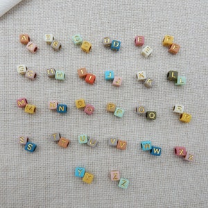 May include: A set of alphabet letter beads in various colors, including pink, blue, green, yellow, and brown. Each bead is a cube with a gold letter on each side.