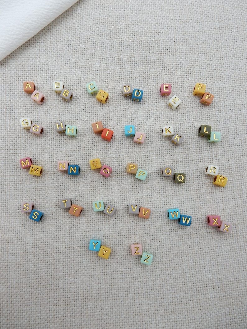 May include: A set of alphabet letter beads in various colors, including pink, blue, green, yellow, and brown. Each bead is a cube with a gold letter on each side.
