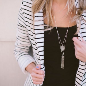 Silver necklace with two layers of chain. The shorter chain hangs a round silver pendant with grooved texture with small black flecks. Longer chain hangs a rectangle pendant with the same grooved texture.