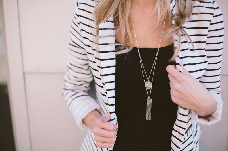 Silver necklace with two layers of chain. The shorter chain hangs a round silver pendant with grooved texture with small black flecks. Longer chain hangs a rectangle pendant with the same grooved texture.