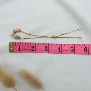 May include: A gold chain bracelet with a sliding clasp. The bracelet has pink, white, and brown beads with the letters "F I M Y" on them. The bracelet is measured on a pink tape measure that reads "1-2-3-4-5-6" inches.