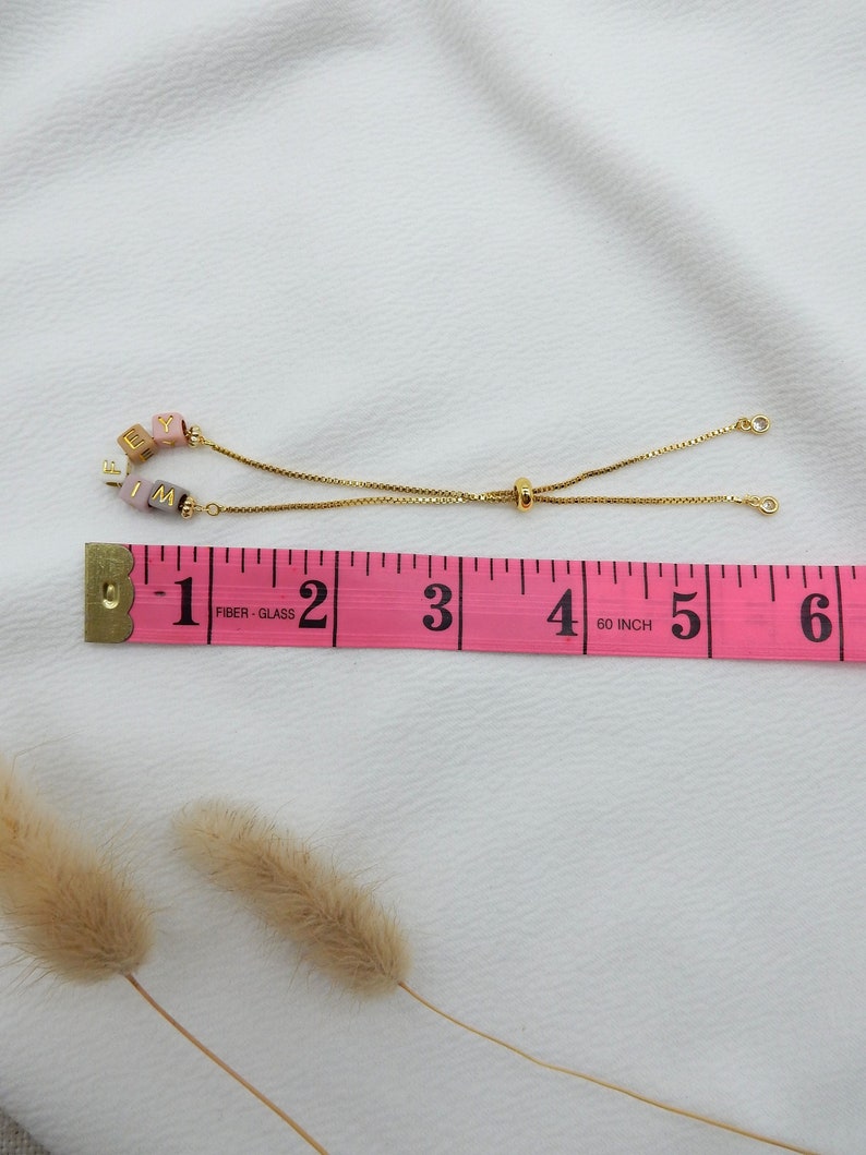 May include: A gold chain bracelet with a sliding clasp. The bracelet has pink, white, and brown beads with the letters "F I M Y" on them. The bracelet is measured on a pink tape measure that reads "1-2-3-4-5-6" inches.