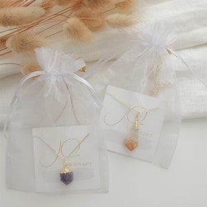 May include: Two necklaces with crystal pendants packaged in white organza bags. The necklaces are on white cards with the word "JO" written in cursive. One necklace has a purple amethyst pendant and the other has a clear and orange citrine pendant.