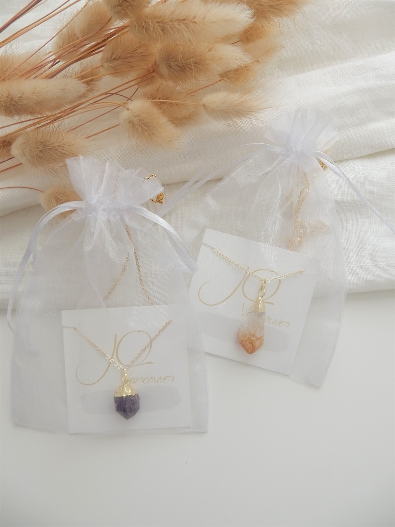 May include: Two necklaces with crystal pendants packaged in white organza bags. The necklaces are on white cards with the word "JO" written in cursive. One necklace has a purple amethyst pendant and the other has a clear and orange citrine pendant.