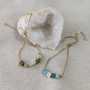 May include: Two gold chain bracelets with adjustable sliding knots. One bracelet has the name "XANDER" spelled out in colorful square beads. The other bracelet has the name "LIAM" spelled out in colorful square beads.
