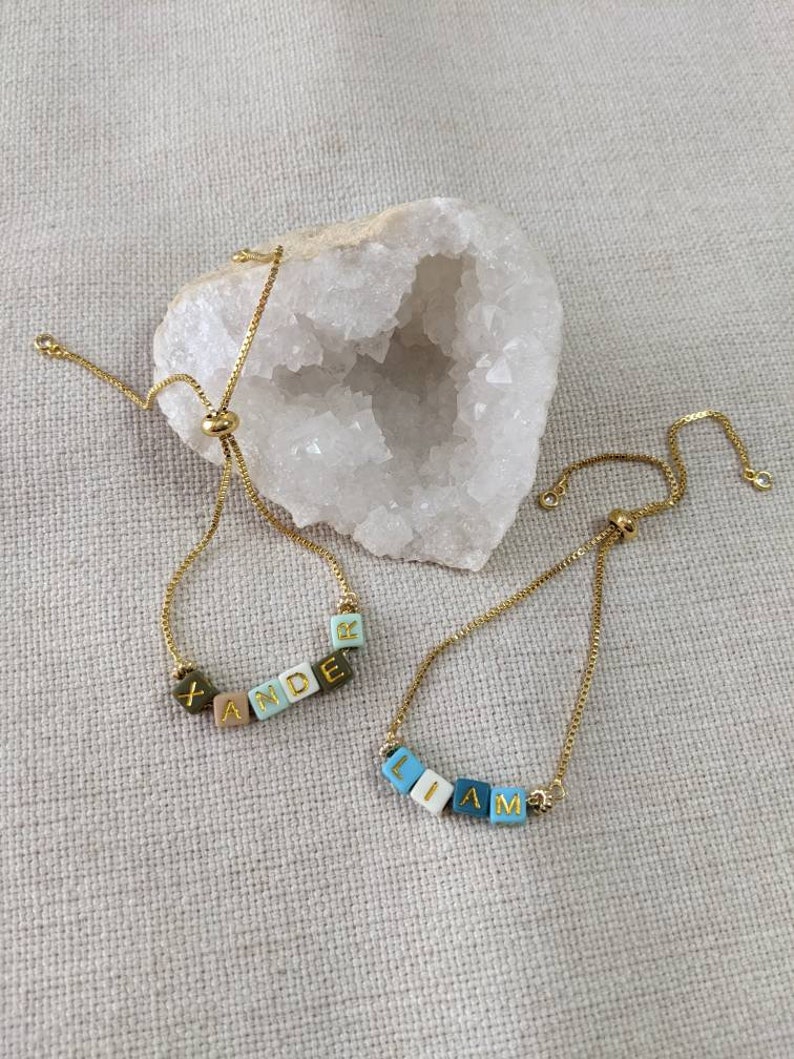 May include: Two gold chain bracelets with adjustable sliding knots. One bracelet has the name "XANDER" spelled out in colorful square beads. The other bracelet has the name "LIAM" spelled out in colorful square beads.