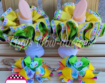 May include: A pair of white ankle socks with yellow, green, and rainbow ribbon ruffles. Two matching hair bows with yellow, green, and white paint splatter ribbon and a white paint palette charm. The text "That's Bow-Dacious!" is on a pink and black circle.
