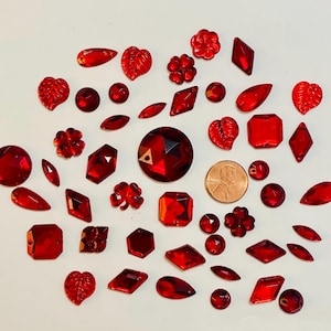 May include: A collection of red acrylic gemstones in various shapes, including round, leaf, flower, and geometric designs. A penny is included for size reference. The gemstones are scattered on a white surface.