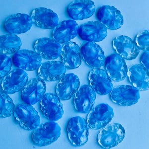 May include: A collection of light blue, oval-shaped cabochons with a faceted design. The cabochons are scattered across a light blue surface, creating a visually appealing arrangement. These decorative pieces could be used for jewelry making or other craft projects.