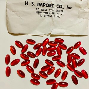May include: A collection of oval, translucent, red capsules scattered on a white surface. A piece of paper with the text "H. S. IMPORT CO., Inc. 20 WEST 37th STREET NEW YORK 18, N. Y. TEL. BRYANT 9-1830" is in the background.