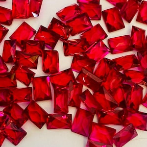 May include: A collection of radiant, rectangular, faceted red gemstones scattered across a white surface. The gems vary slightly in size and shape, catching the light and displaying a vibrant, ruby-like color. Ideal for jewelry making or craft projects.