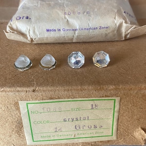 May include: Four clear crystal rhinestones of varying shapes and sizes are displayed on a brown surface. A paper package is visible in the background, with text that reads "Made in Germany (American Zone)." A green label reads "crystal" and "Gross."