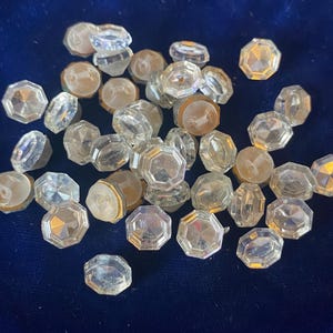 May include: A collection of clear, faceted glass buttons with a variety of shapes and sizes. Some buttons have a flat back, while others have a rounded back. The buttons are scattered on a dark blue surface.