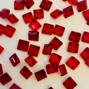 May include: Scattered across a white surface are numerous small, square, faceted, ruby-red gemstones. The gems have a brilliant, transparent appearance, catching the light and reflecting it. The overall effect is one of sparkle and luxury.