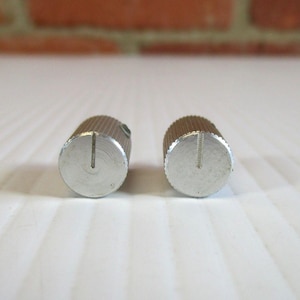 May include: Two silver-colored control knobs with a grooved cylindrical body and a flat, circular top. Each knob has a vertical groove cut into the top. The knobs are set against a white surface with a brick background.