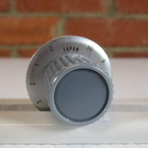 Puede incluir: Un mando de control gris con un dial circular y un agarre estriado. El dial está marcado con números del 0 al 10 y la palabra "JAPAN". El mando está sobre una superficie blanca y un fondo de ladrillo.
